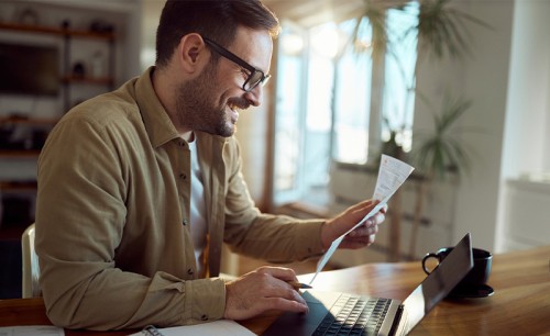 Young happy man analyzing his bills while paying them over laptop at home