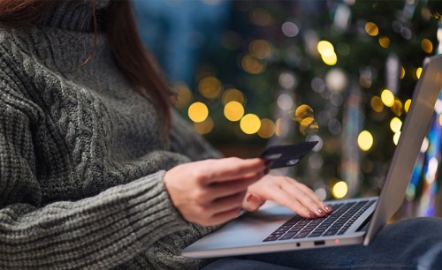 Woman using her credit card to make an online purchase on her laptop, with Christmas lights in the background.