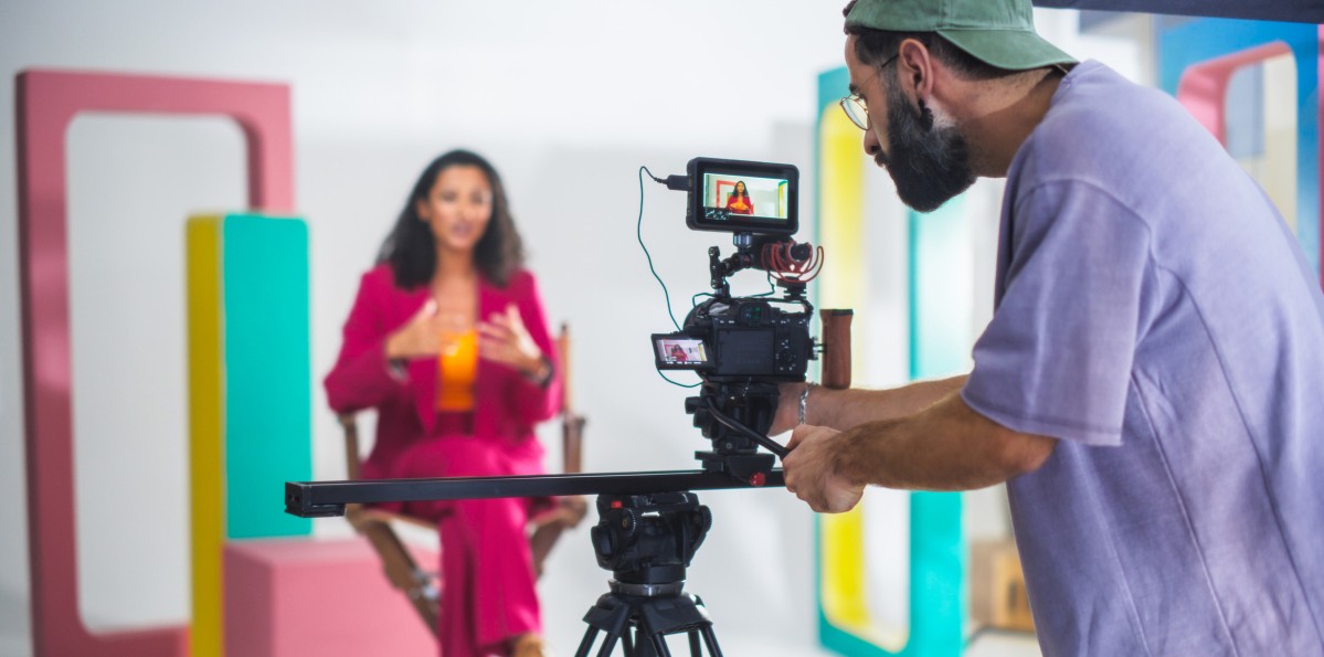 Young Male Cinematographer With Beard, Wearing Cap, Records Vibrant Female Host In Pink Suit During Lively Talk Show Set With Colorful Geometric Shapes