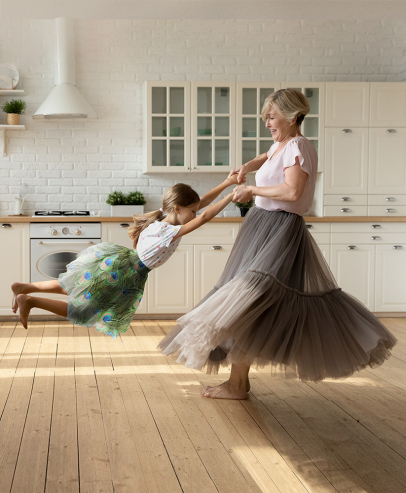 Grandmother and granddaughter dancing in a bright kitchen wearing flowing tulle skirts. The granddaughter's skirt has a peacock feather pattern.